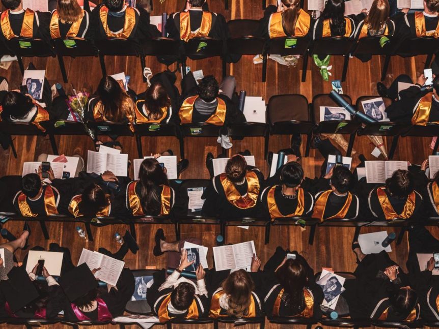 An aerial view of rows of seated graduation students with colourful sashes over their black academic robes.